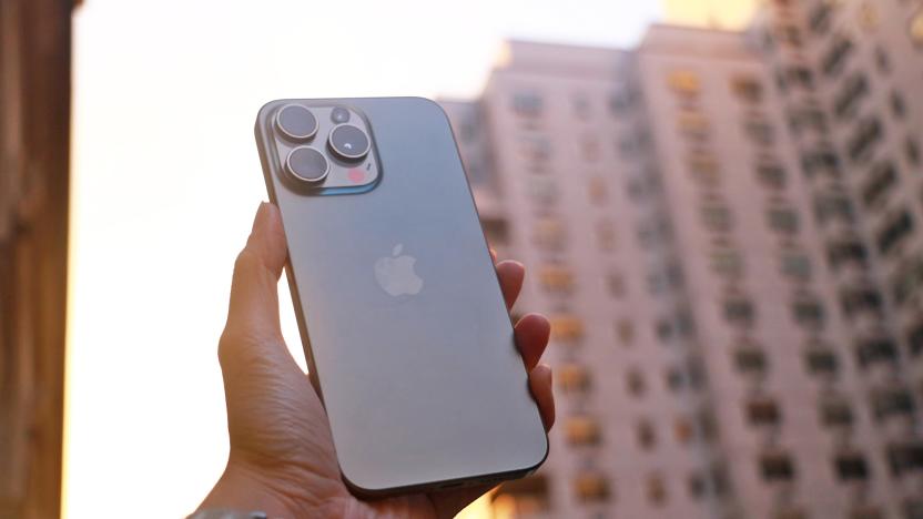 The iPhone 15 Pro Max held up in mid-air with a New York building in the background while the sun sets.   