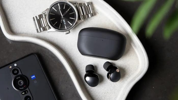 Sony WF-1000XM5 earbuds in black pictured on a tray next to the charging case, a silver watch and a Sony smartphone