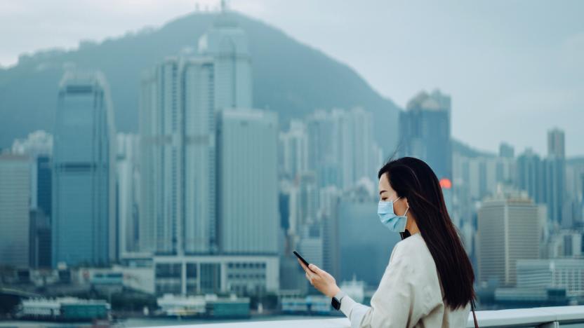 Young Asian woman with surgical face mask   to prevent the spread of viruses during the Covid-19 health crisis and using smartphone against the iconic city skyline of Hong Kong