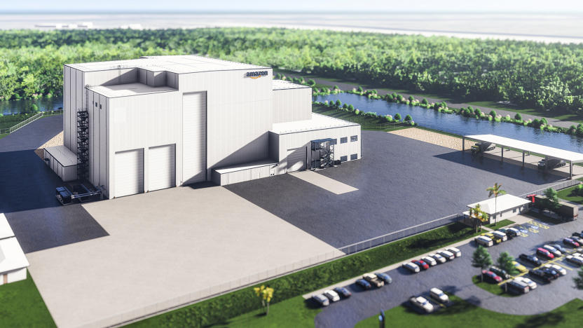 Rendered photo of Amazon’s satellite-processing facility for its Project Kuiper satellite broadband initiative. A large light-colored building with an Amazon logo sits next to a large lot. It’s surrounded by a parking lot (full of cars) with water and trees in the background.