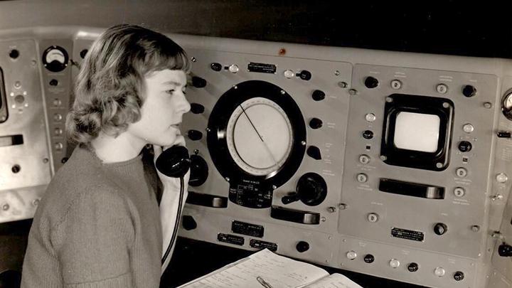 Archival black & white photo of Virginia Norwood, sitting in a (mid-20th-century) control room while talking on the phone and jotting notes in a log book.