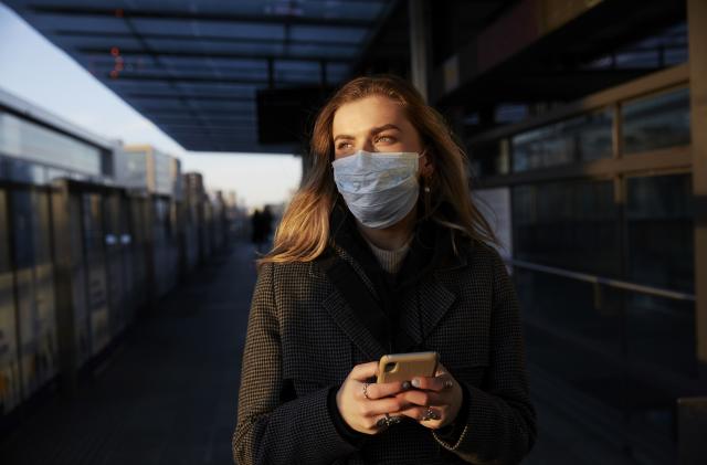 Young woman standing on train station wearing protective mask, using phone