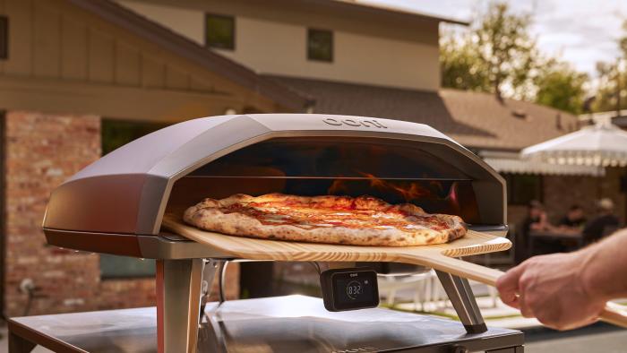 A person's hand using a pizza peel to remove a large cooked pizza from an outdoor pizza oven that's located on a patio.