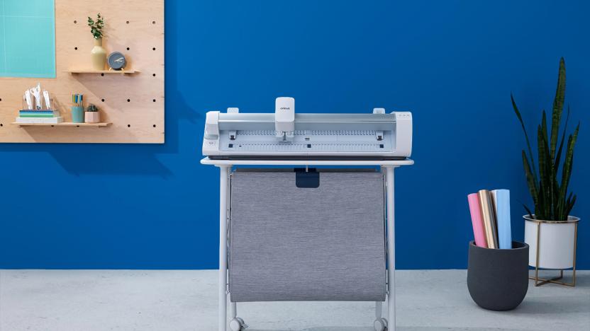 Marketing photo of the Cricut Venture, a consumer and small-business cutting machine for craft projects. It sits on a stand with a drape of fabric hanging below. It sits in a stylish room with a blue wall, gray floor and modern motif.