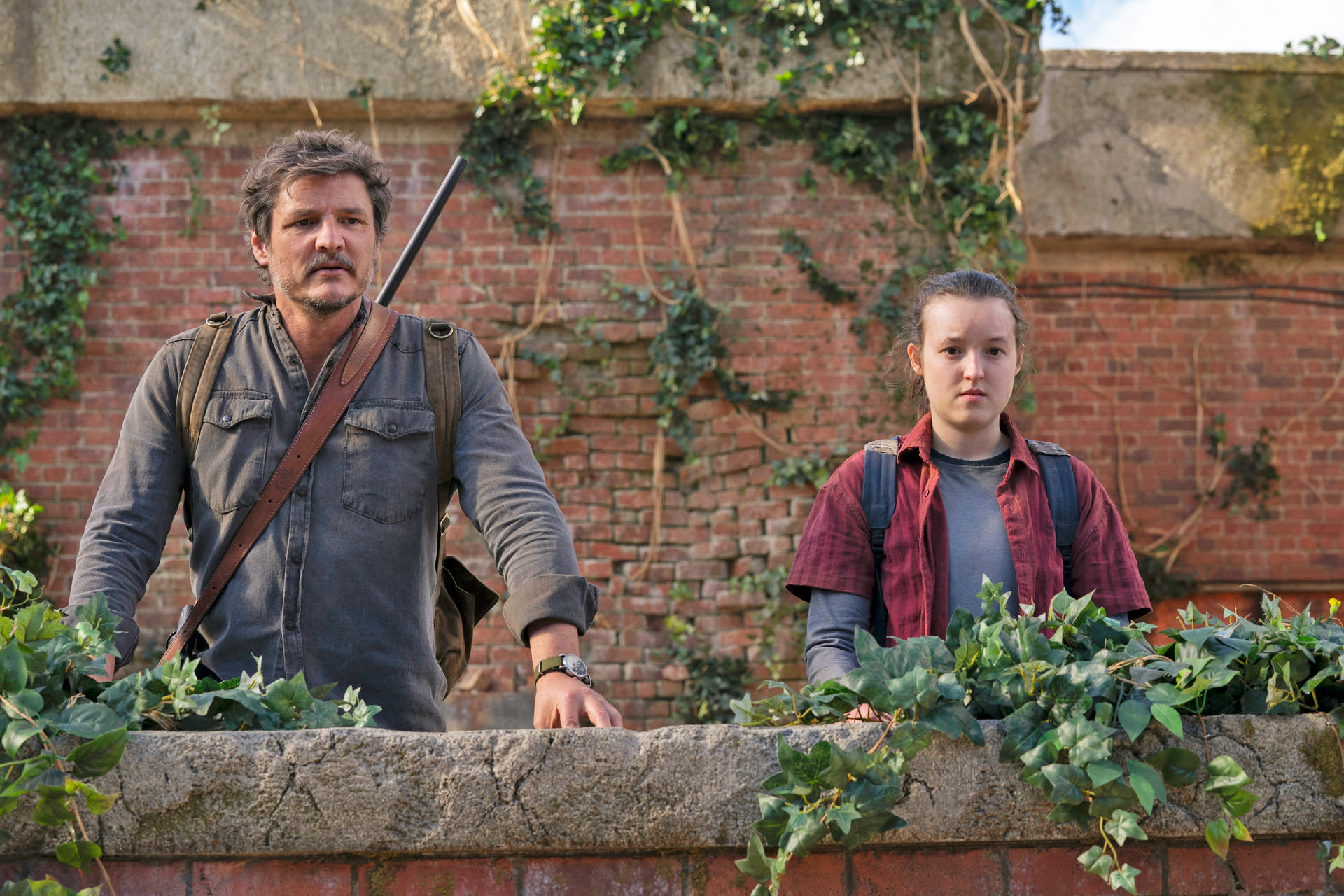 Pedro Pascal and Bella Ramsey stand behind a short brick wall and look at the camera in this promotional image from the TV series 'The Last of Us'.