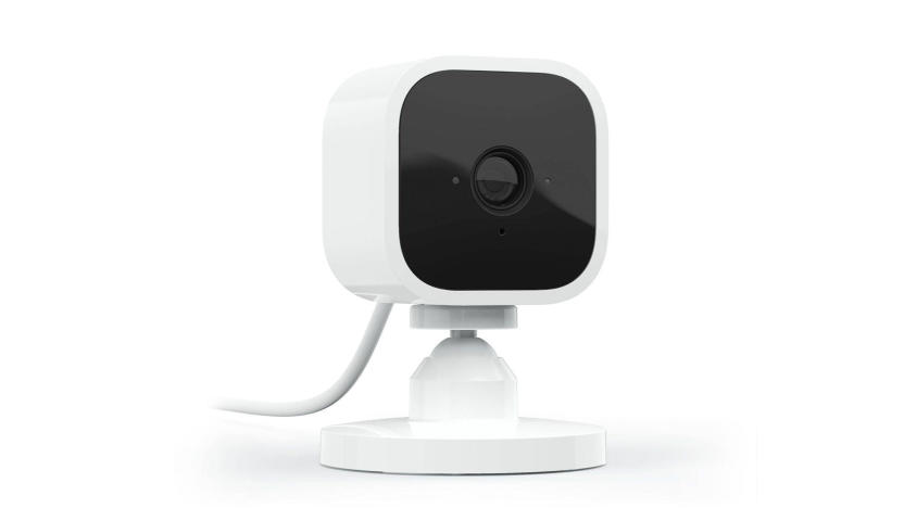 Marketing photo for the Blink Mini indoor security camera. The camera has a black front face and a white body. It’s facing the right with a power cord fading off to the left.