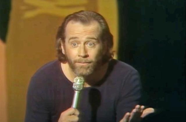 George Carlin holds a microphone and eyeballs the crowd in a questioning manner in the middle of doing a bit on stage in the 1970s.