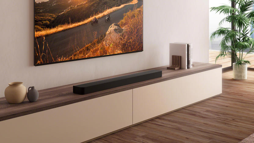 A black soundbar on a wooden shelf underneath a TV with a mountain road sunset scene on the display.