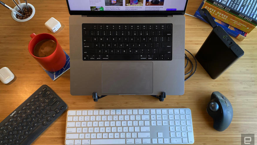 A MacBook Pro sits on a stand on a desk, surrounded by various keyboards and other useful accessories. 