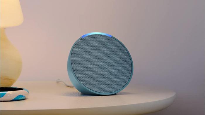 The Amazon Echo Pop speaker in blue sits on a night table with a yellow lamp nearby. 