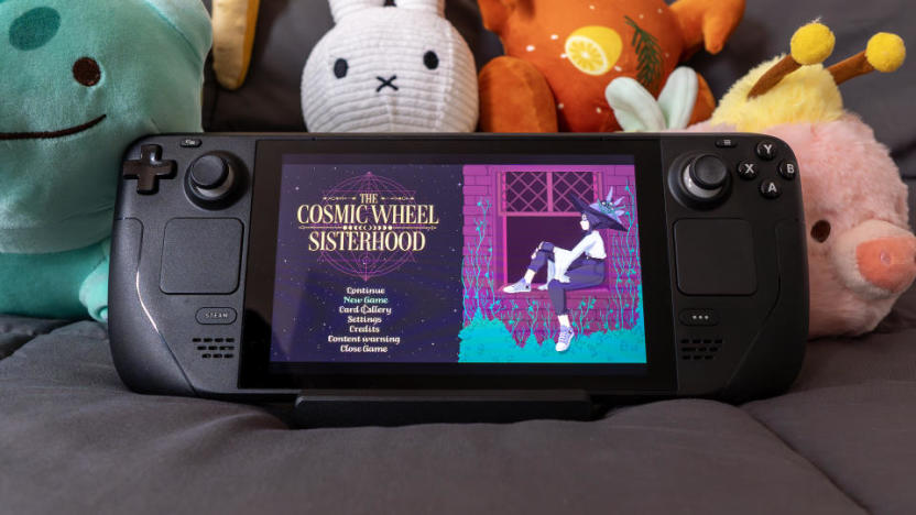 The Steam Deck sitting on a blanket with cute plush toys behind it. The Cosmic Wheel Sisterhood title screen is showing on its display.