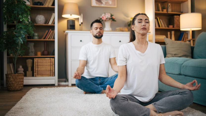 Picture of a man and a woman sitting on the floor in a modern apartment and practicing yoga. They wear white shirts.