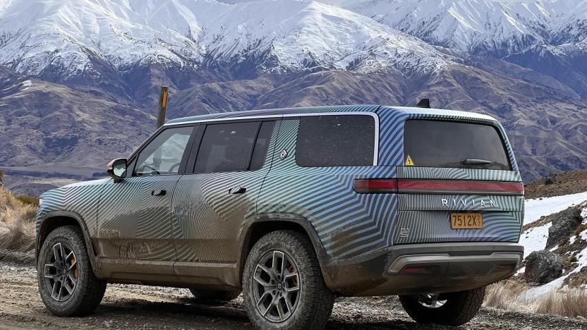 A Rivian R1S SUV on a rocky and muddy road with snow capped mountains in the background.