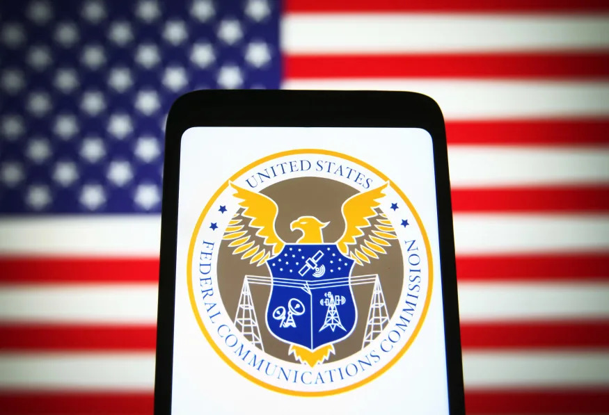 UKRAINE - 2021/11/25: In this photo illustration, U.S. Federal Communications Commission (FCC) seal is seen on a smartphone screen with the US flag in the background. (Photo Illustration by Pavlo Gonchar/SOPA Images/LightRocket via Getty Images)