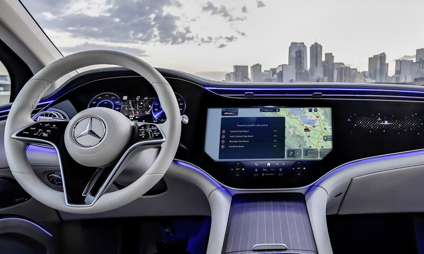 An in-car view of a modern Mercedes interior. The steering wheel is on the left, with main console in the center. In the background is a city skyline.