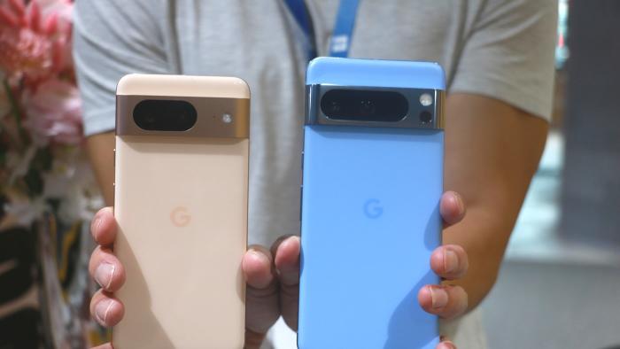 A person holding up the peach Pixel 8 and the blue Pixel 8 Pro with their rear cameras facing out.