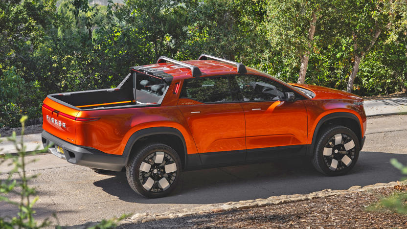 A Fisker Alaska 2023 pickup in burnt orange color is seen driving along a wooded road on a hillside, with trees flanking the road.