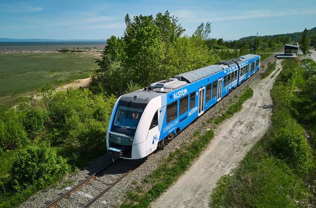 View from slightly above of the Coradia iLint passenger train (blue and white, two cars) moving along a lush green Canadian countryside.