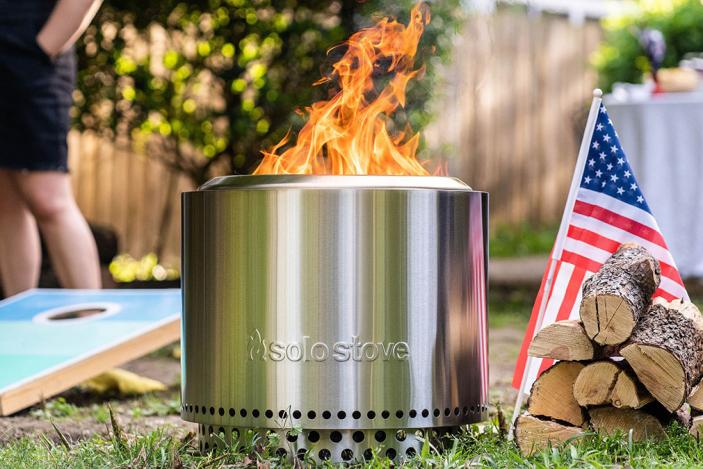 A Solo Stove fire pit sitting on grass next to fire wood and an American flag at a backyard party.