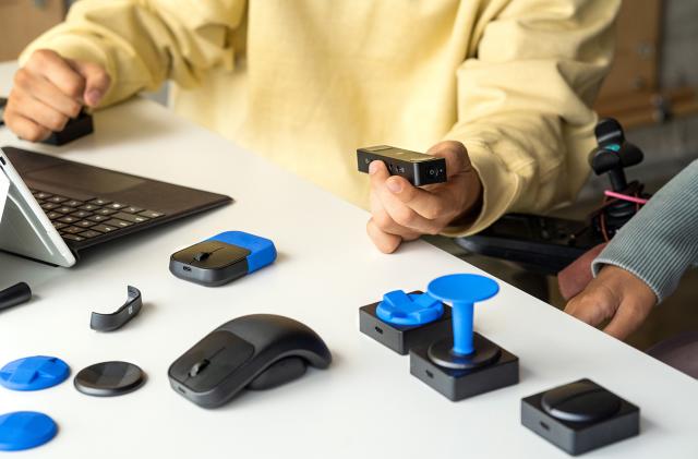 The Microsoft Adaptive Mouse on a table, docked onto a curved fin and surrounded by other inputs and blue parts making different controllers.