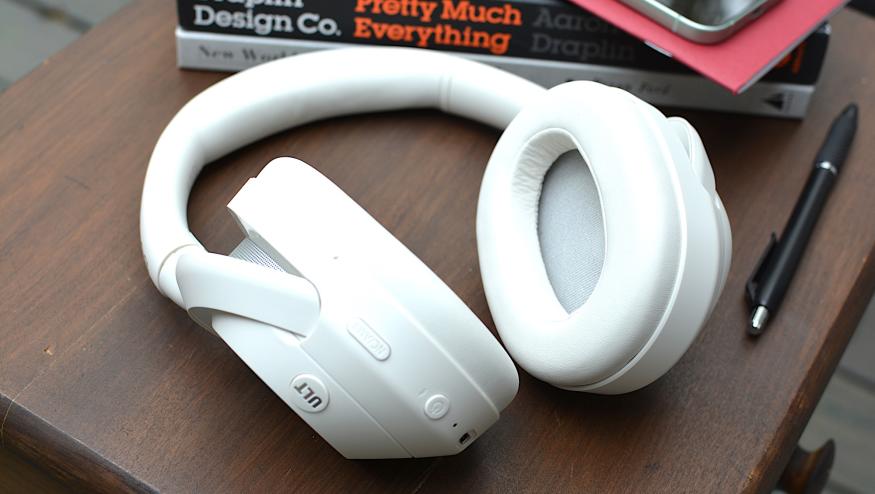 White headphones sitting on a wooden table from above with a red notebook and phone in the background sitting on top of a black and a white book.