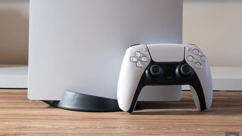 Closeup of a DualSense controller leaning against the PS5 console. Only the bottom part of the (upright) console is visible. Wood-grained desk with blurred background.