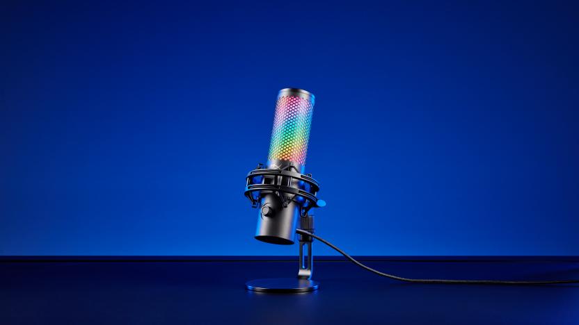 Product image of the HyperX QuadCast 2 S gaming mic. The mic (with over 100 LEDs) sits on a black desk in front of a blue background. Dramatic shadows.