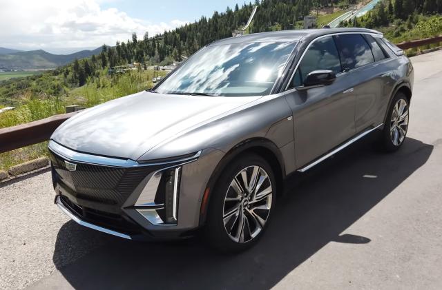 A gray Cadillac Lyriq driving on a hillside road with green trees in the background. Photo taken from the front left of the vehicle.