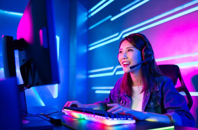 Young woman wearing a headset while sitting at a computer. The keyboard and backdrop are lit in neon colors.