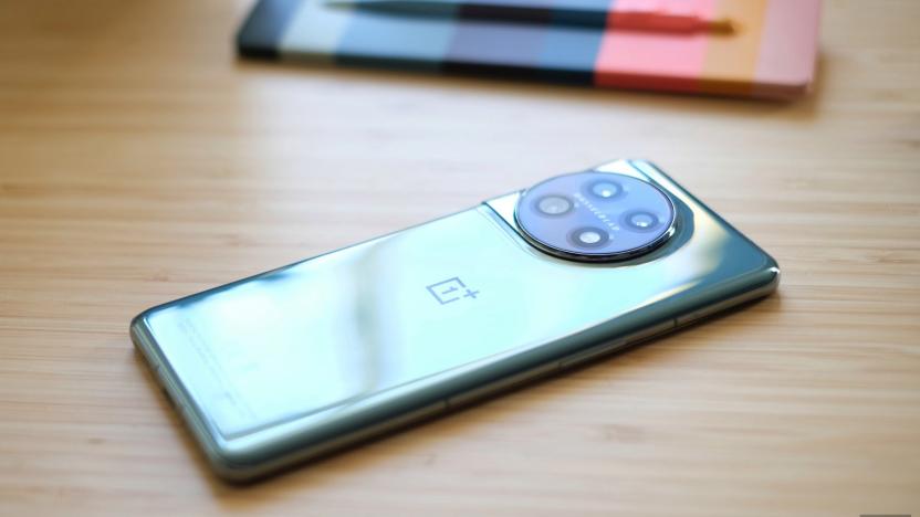 A green metallic OnePlus 11 5G smartphone sits face-down on a wooden table top with a pen and notepad in the background.