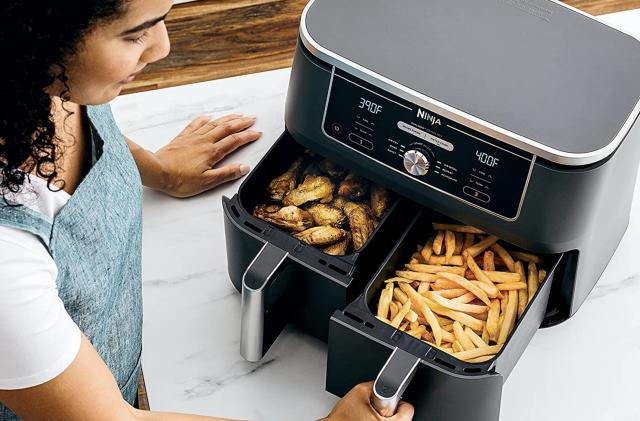A woman uses the dual-zone Ninja DZ401 Foodi air fryer on a white marble kitchen countertop. There are french fries in one compartment and chicken wings in the other. 