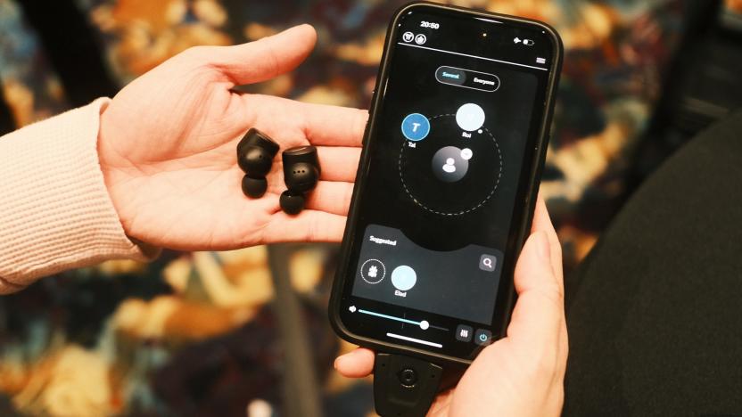 A pair of black earbuds on a person's palm, with their other hand holding up a phone with the OrCam Hear app on its screen.