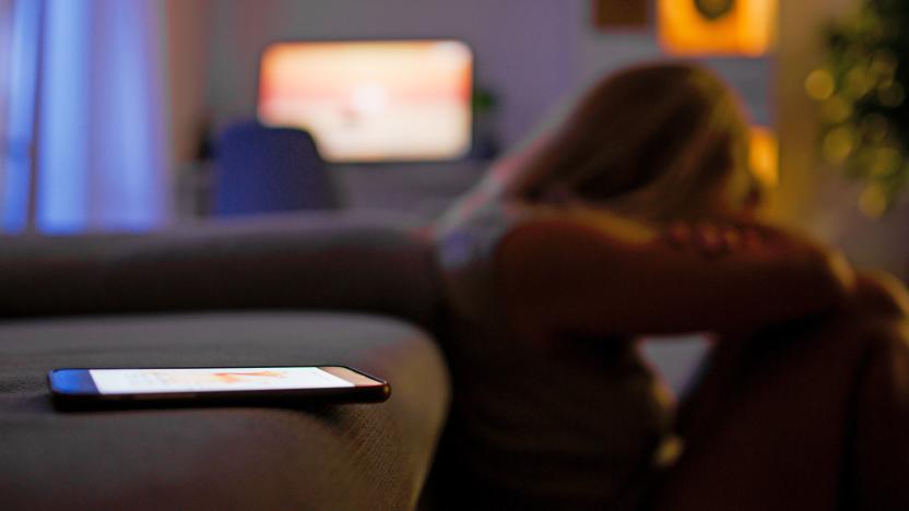 A seemingly depressed woman sits on floor in front of a couch hugging her knews, with a mobile phone in the foreground on the sofa.