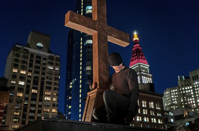 Daredevil looks down on the city from his perch holding onto a cross at the top of a church.