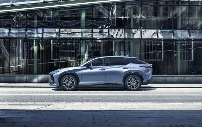 The Lexus RZ sport sedan in light metallic blue parked in front of a modern office building that's mostly made of glass windows. 