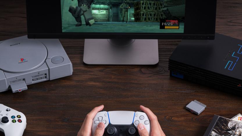 A pair of hands holds a PlayStation 5 DualSense controller. A PlayStation 1 and PlayStation 2 are in the background and a dongle sits on the table. A game is shown on a monitor.