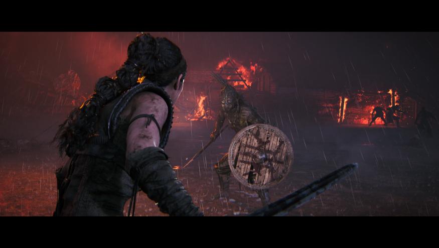 Dark setting, with the protagonist Senua wielding a sword in the foreground. A grotesque enemy with a shield lies ahead with burning buildings in the background.