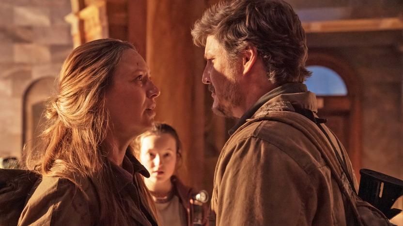 Tess (Anna Torv) and Joel (Pedro Pascal) stand face-to-face with serious expressions in The Last of Us. Ellie (Bella Ramsay) is slightly out of focus in the background.