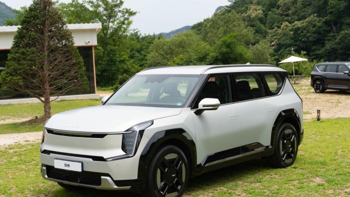 A white Kia EV9 SUV is parked in a grassy park area with woods in the background.