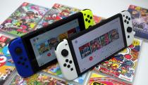 Two Nintendo Switch consoles sitting upright on an array of game boxes. Standard model with blue and yellow Joy-Cons on the left, OLED model with white Joy-Cons on the right.