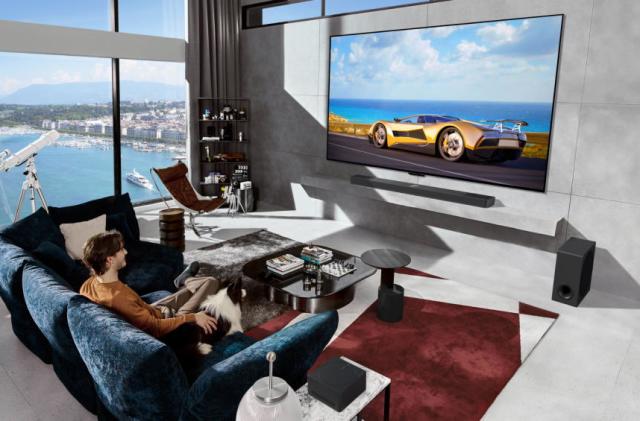 A man sits on a blue couch with his dog watching a race car on his humongous LG TV. It's a modern bachelor pad overlooking a bay with a telescope by the large windows.