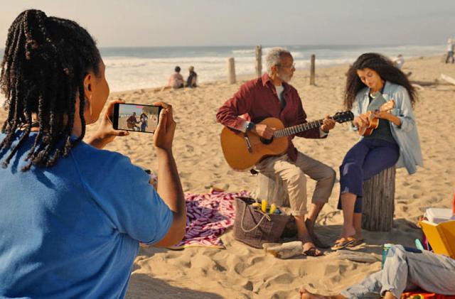 A person holds an iPhone to capture spatial video of an older man and younger woman strumming stringed instruments on the beach.