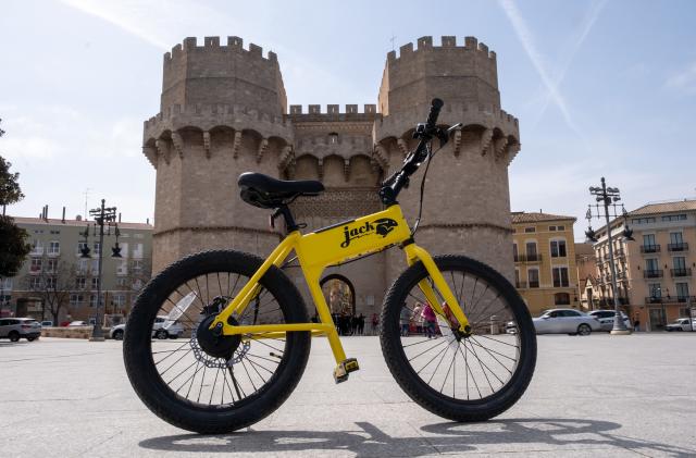 The JackRabbit ebike in yellow on its own in an empty plaza ini front of a small castle-like structure with two turrets.