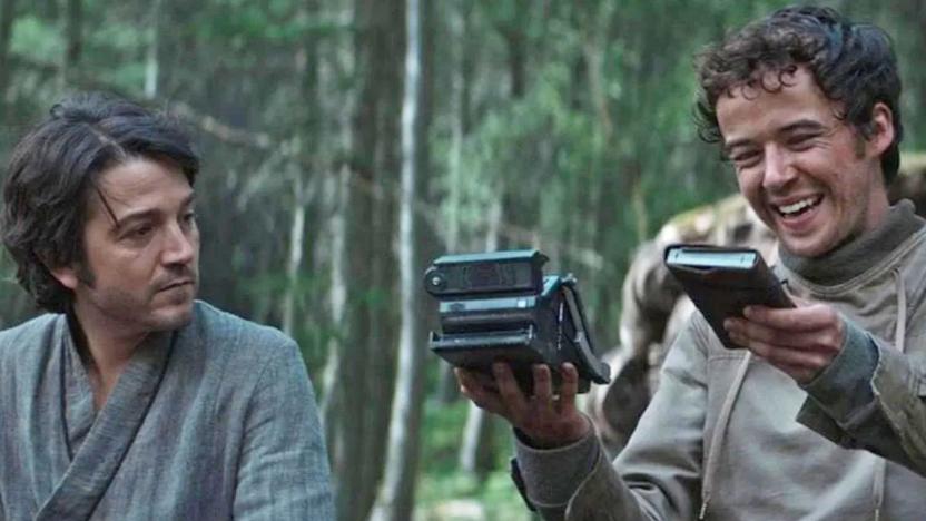 A still image from the tv series 'Andor' showing the main character sitting next to someone who's holding a device that appears to look like a folding polaroid camera. 