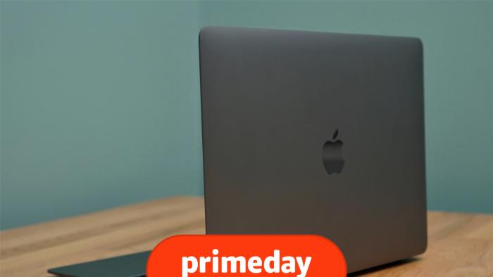 A silver MacBook Air laptop sits on a brown desk in front of a blue wall, with its back facing the camera at an angle, showcasing an Apple logo.