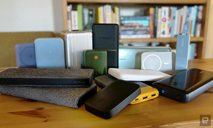 Fourteen different power banks and battery packs from brands like Anker, Otterbox, and Mophie arranged on a wooden table with a bookcase in the background. 