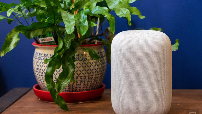Google Nest Audio smart speaker sitting on a wooden tablet next to a green plant, against a blue wall.
