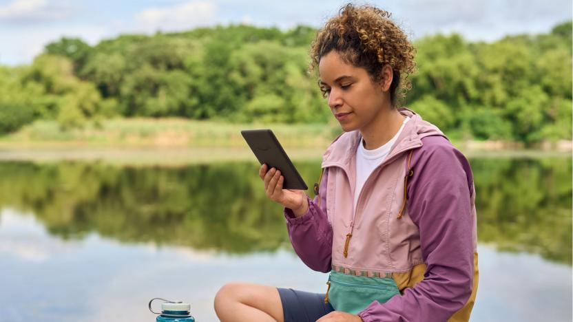 A woman sits on a rock near a lake reading from an Amazon Kindle