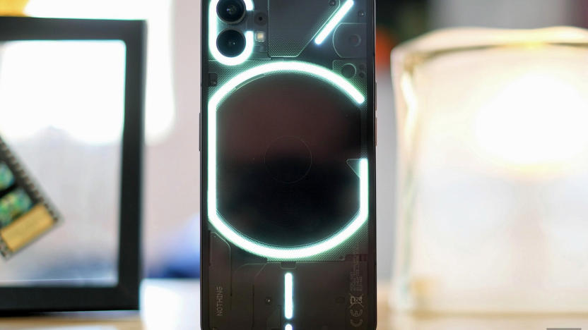 The new Nothing Phone 1 stands on its end on a table. The phone is black with a see-through back showing a glowing series of lines and rings.
