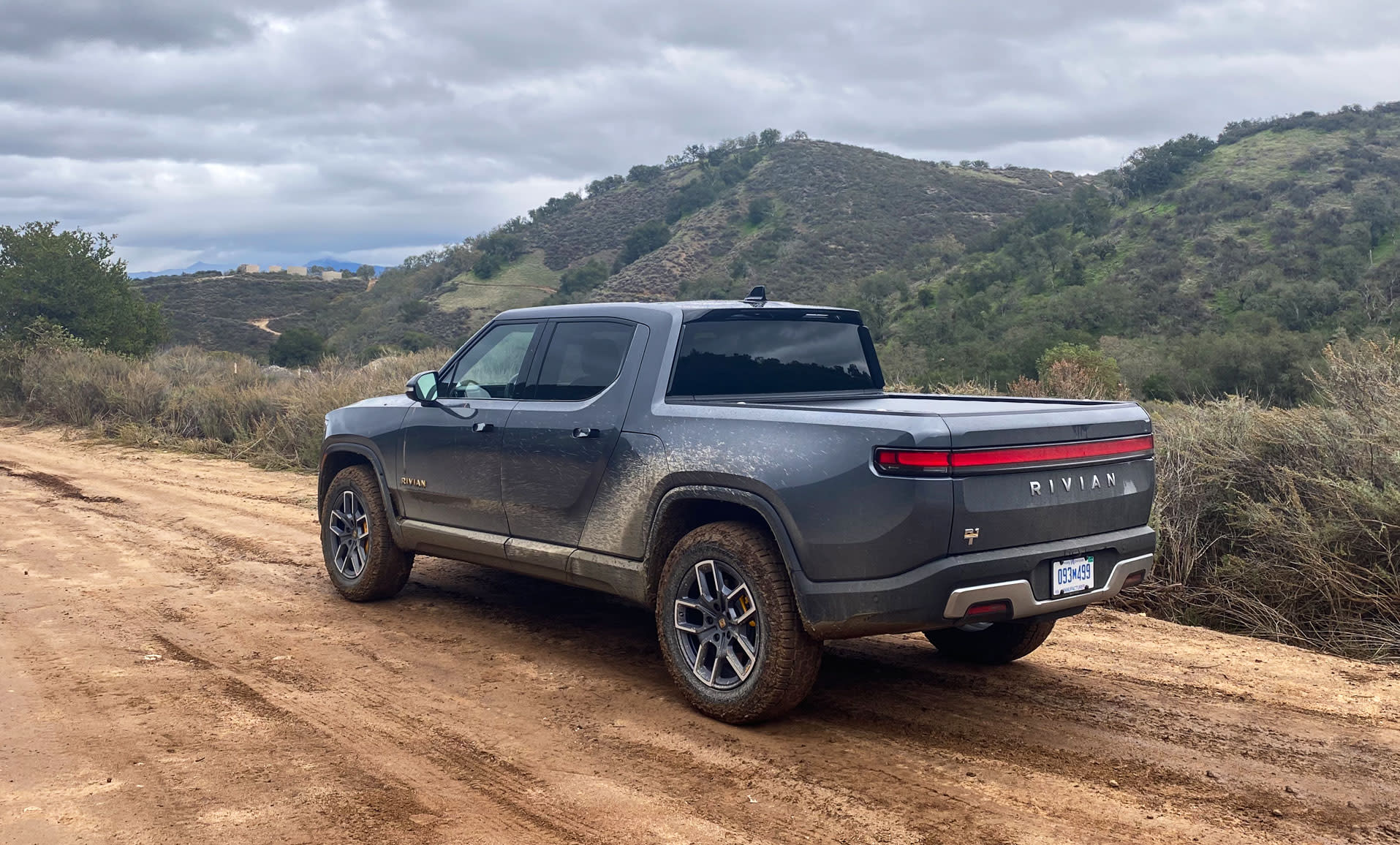 A dark grey Rivian R1T drives up a dirt road that's headed upwards with hills in the background.